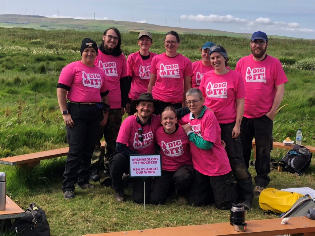 A group of people in an outdoor setting wearing pink Dig It! t-shirts and holding a sign that says: Archaeology in Action - Ask Us About Our Work
