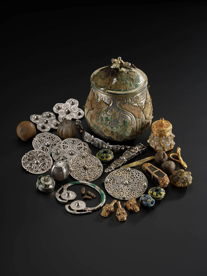 The Galloway Hoard with the silver vessel wrapped in plant fibre and wool textiles and various other items around it, including brooches, beads and dirt balls