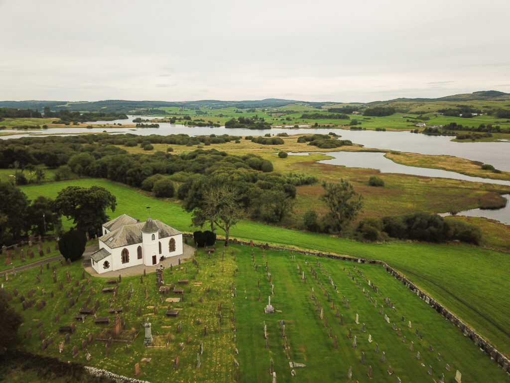 Aerial photo of a rural Scottish setting with waterlogged plains in the background and a white church and graveyard in the foreground
