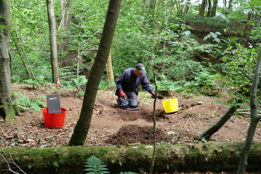 Photo of a person in a sweatshirt and dark cap kneeling by an archaeological pit and spooning dirt into a yellow bucket