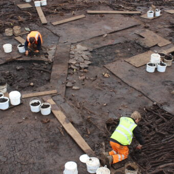 Aerial photo of three archaeologists in orange and yellow high-vis clothing kneeling on wooden planks while excavating a waterlogged prehistoric roundhouse site