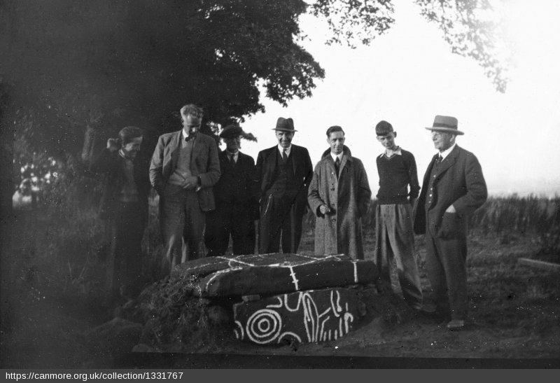 Black and white photo from the early 20th century of a small group of men and boys standing round a prehistoric cist (or coffin) marked with chalk 
