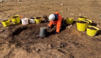 Photo of a person in a white hard hat and orange coat digging in a shallow archaeological trench
