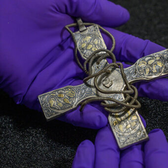 Pre-conservation image taken June 2018 of an ornamented silver pectoral cross with wire chain. The cross is decorated with what appears to be gilding and niello, each arm ornamented with a motif and there appears to be an ornamentation missing from the centre. A chain of coiled silver wire is wrapped around the arms. The cross is from the Viking age Galloway Hoard.