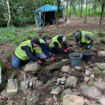 Photo of four people in high visibility clothing kneeling in a shallow archaeological trench in the woods