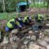 Photo of four people in high visibility clothing kneeling in a shallow archaeological trench in the woods