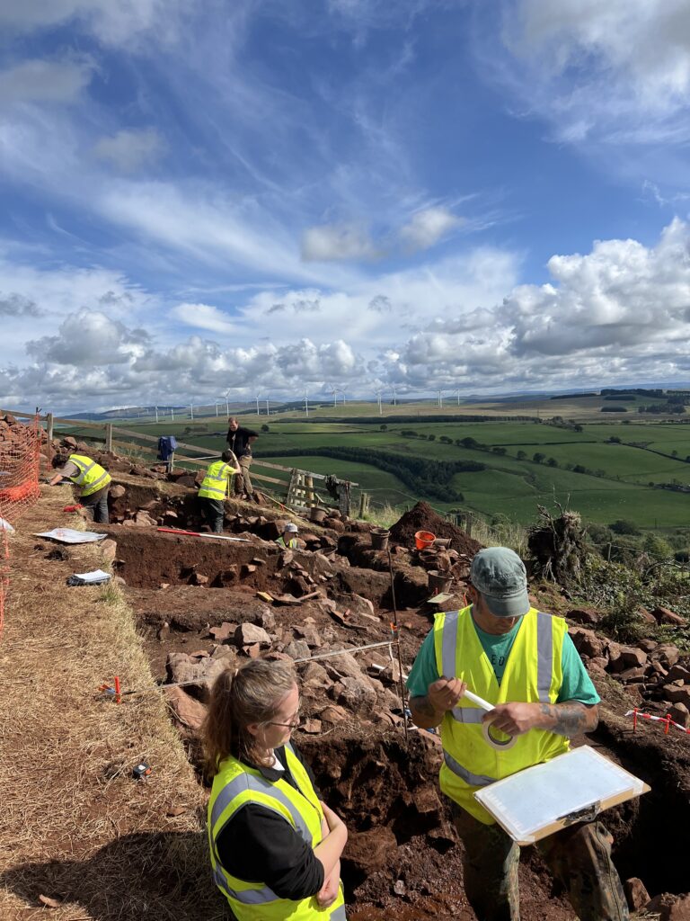 A trench over the rampart of the hillfort, with students beginning to record the sections of the excavation trench. The photograph is taken on the hillfort rampart looking northeast and showcases the extensive views from the site