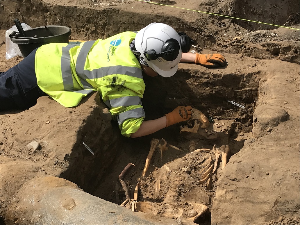 Photo of an archaeologist in a hard hat and high-vis jacket lying over an excavation trench revealing a human burial