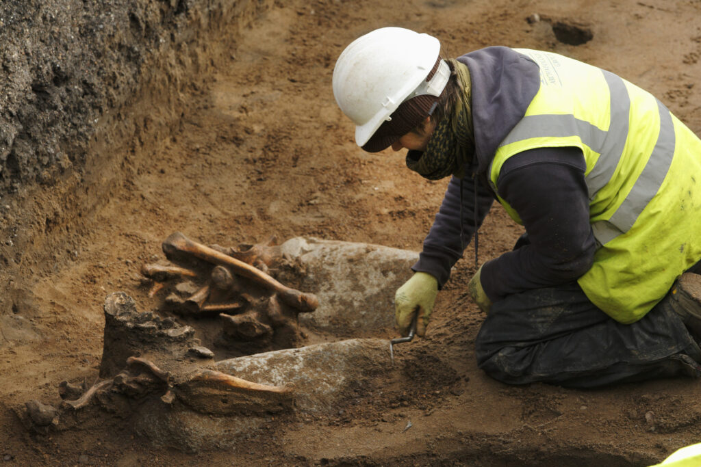 Photo of a person in high-visibility clothing and a white hard hat kneeling in an archaeological trench using a small trowel to excavate human remains