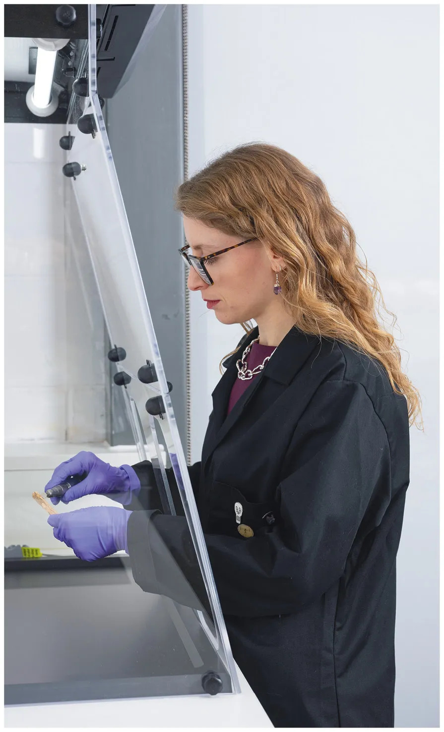 Photo of a museum curator wearing lab clothing sampling a small piece of human bone behind glass