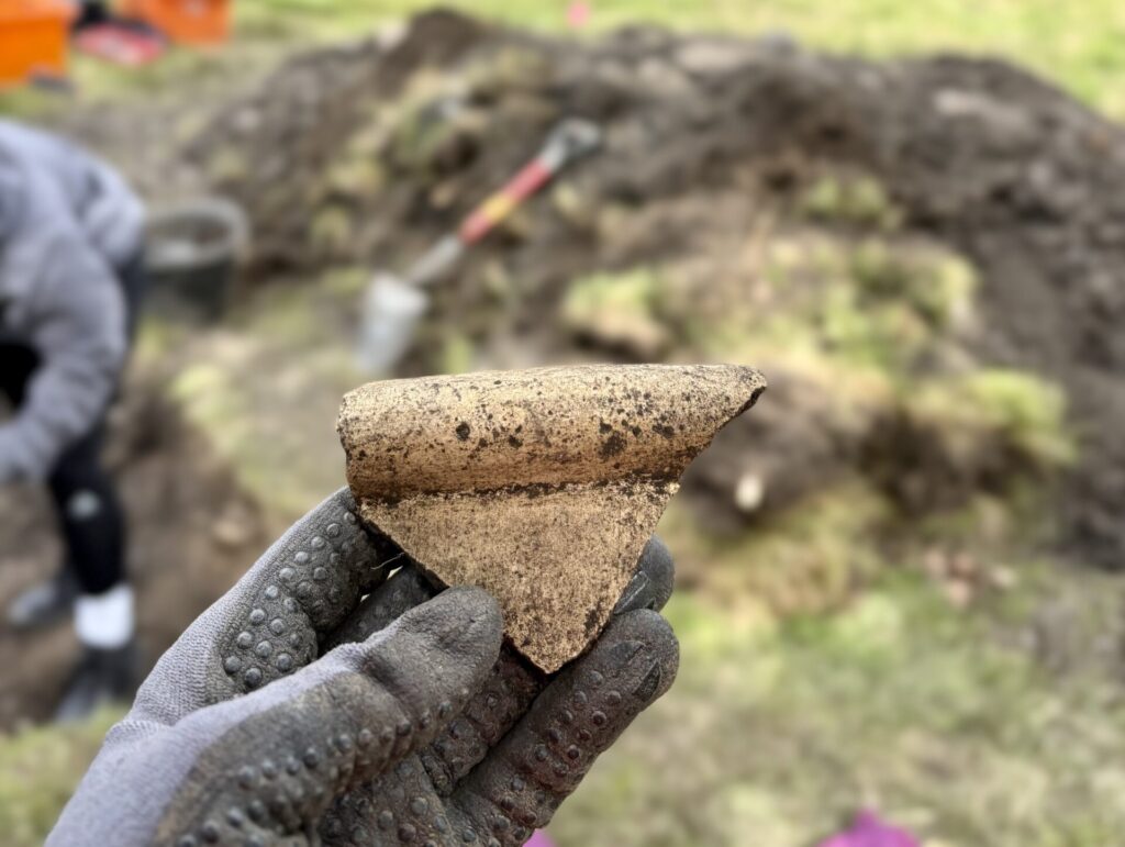 Photo of a gloved hand holding up a triangle-shaped piece of pottery in front of an archaeological trench