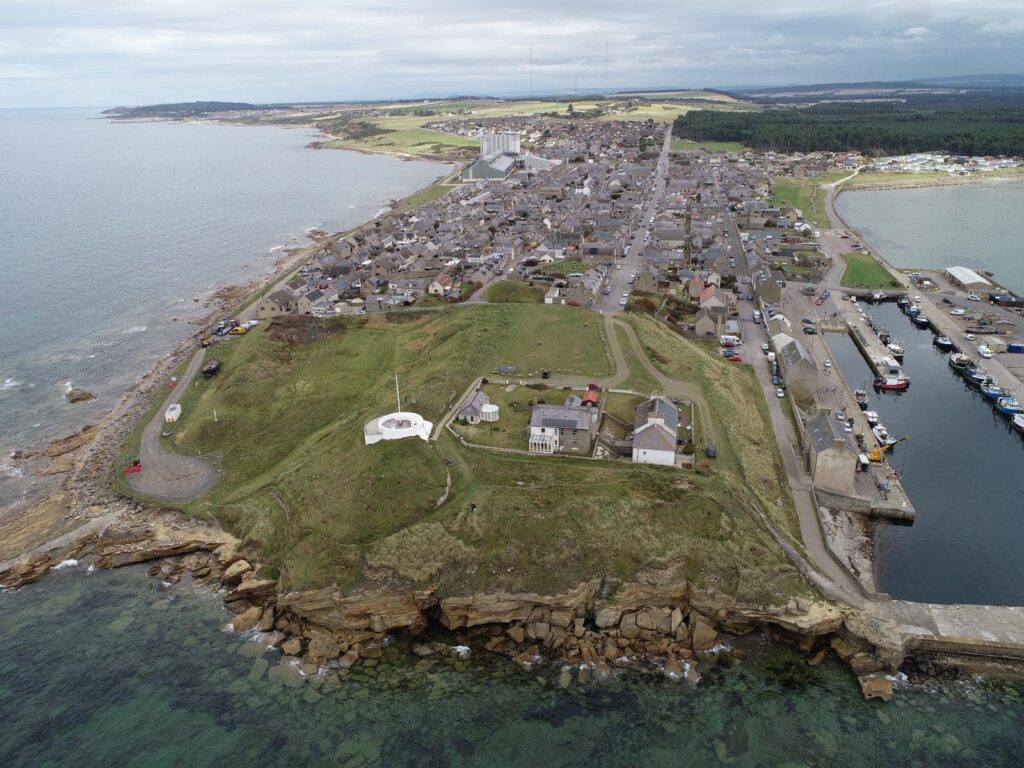 Aerial photograph showing a promontory surrounded by the sea, with a grassy hill area at the end of the promontory and a modern town further back