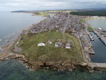 Aerial photograph showing a promontory surrounded by the sea, with a grassy hill area at the end of the promontory and a modern town further back