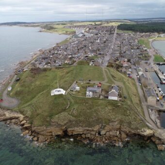 Aerial photograph showing a promontory surrounded by the sea, with a grassy hill area at the end of the promontory and a modern town further back