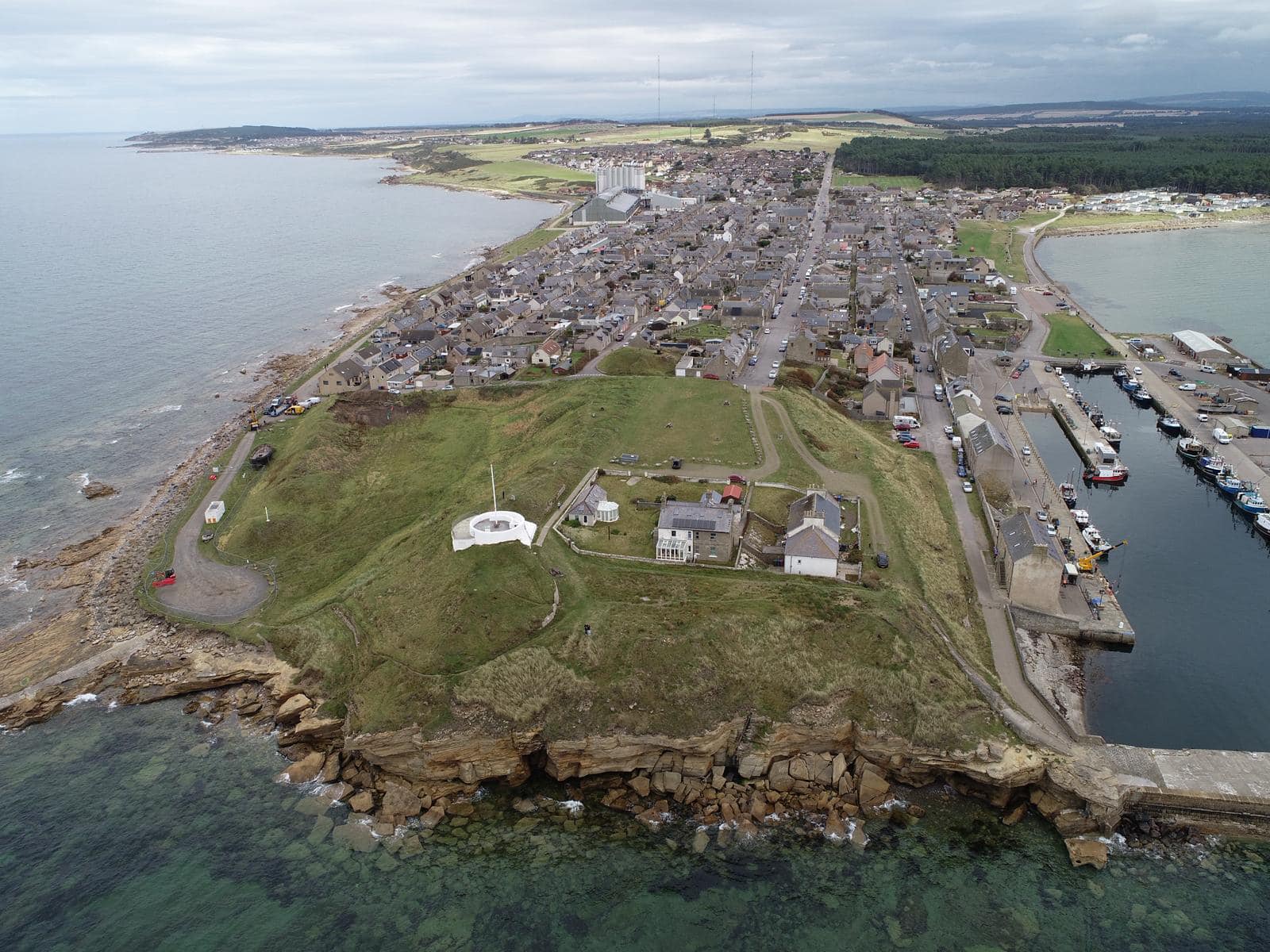 Aerial photograph showing a promontory surrounded by the sea, with a grassy hill area at the end of the promontory and a modern town further back