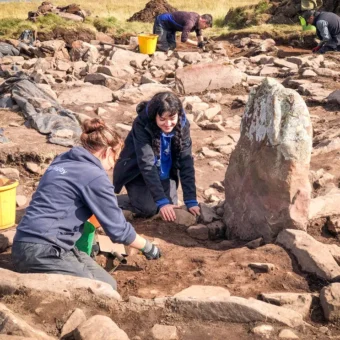 Photo of four people kneeling on the ground of an excavation trench and using trowels to dig