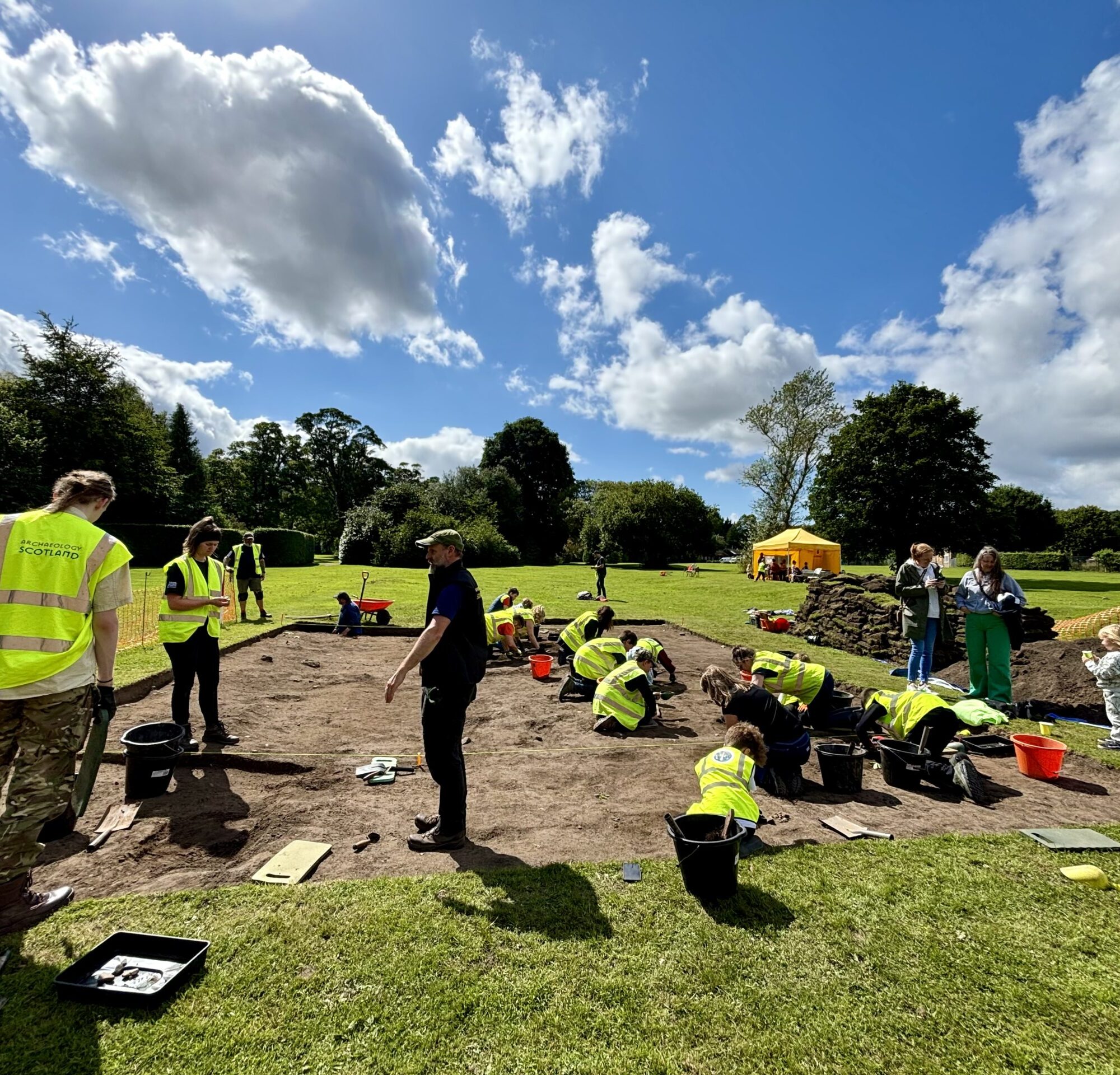 A group of people in high-vis vests digging in a shallow archaeological trench in the sun