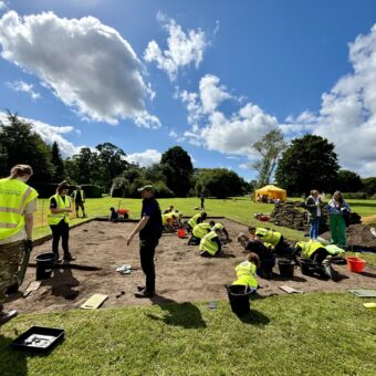 A group of people in high-vis vests digging in a shallow archaeological trench in the sun