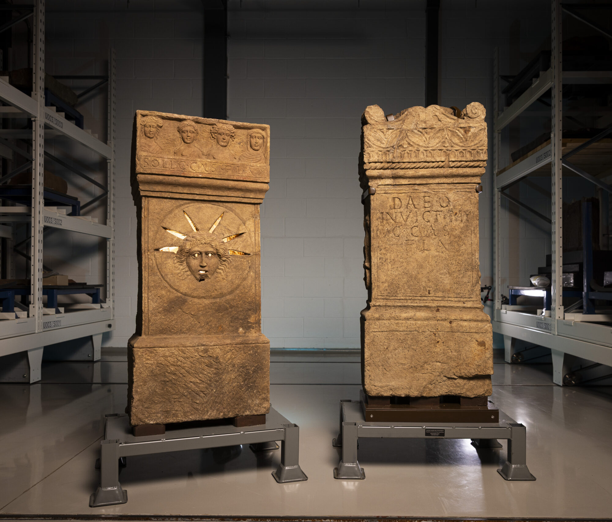 Two Roman altars in a collection centre: One depicts the face of the sun god Sol and would have been illuminated from behind so that in the low light of the temple, worshippers would have seen his eyes, mouth and the rays of his solar crown glowing. It also features carvings of the four seasons - depicted as female deities - conveying the passage of time, another prominent theme within the cult of Mithras. The second honours Mithras himself and is adorned with carvings linked to the god Apollo, another god of light – a griffin, lyre and plectrum - as well as two ravens, which are often seen in Mithraic imagery.