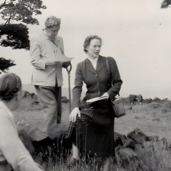 Photo of a woman in 1950s clothing sitting on a low stone wall with people around her
