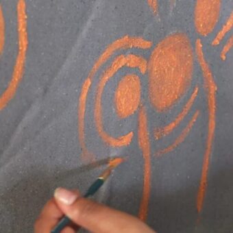 Photo of a hand using a paint brush to paint orange swirls on a grey canvas