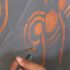 Photo of a hand using a paint brush to paint orange swirls on a grey canvas