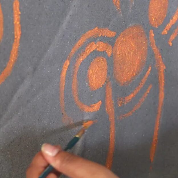 Photo of a hand using a paint brush to paint orange swirls on a grey canvas