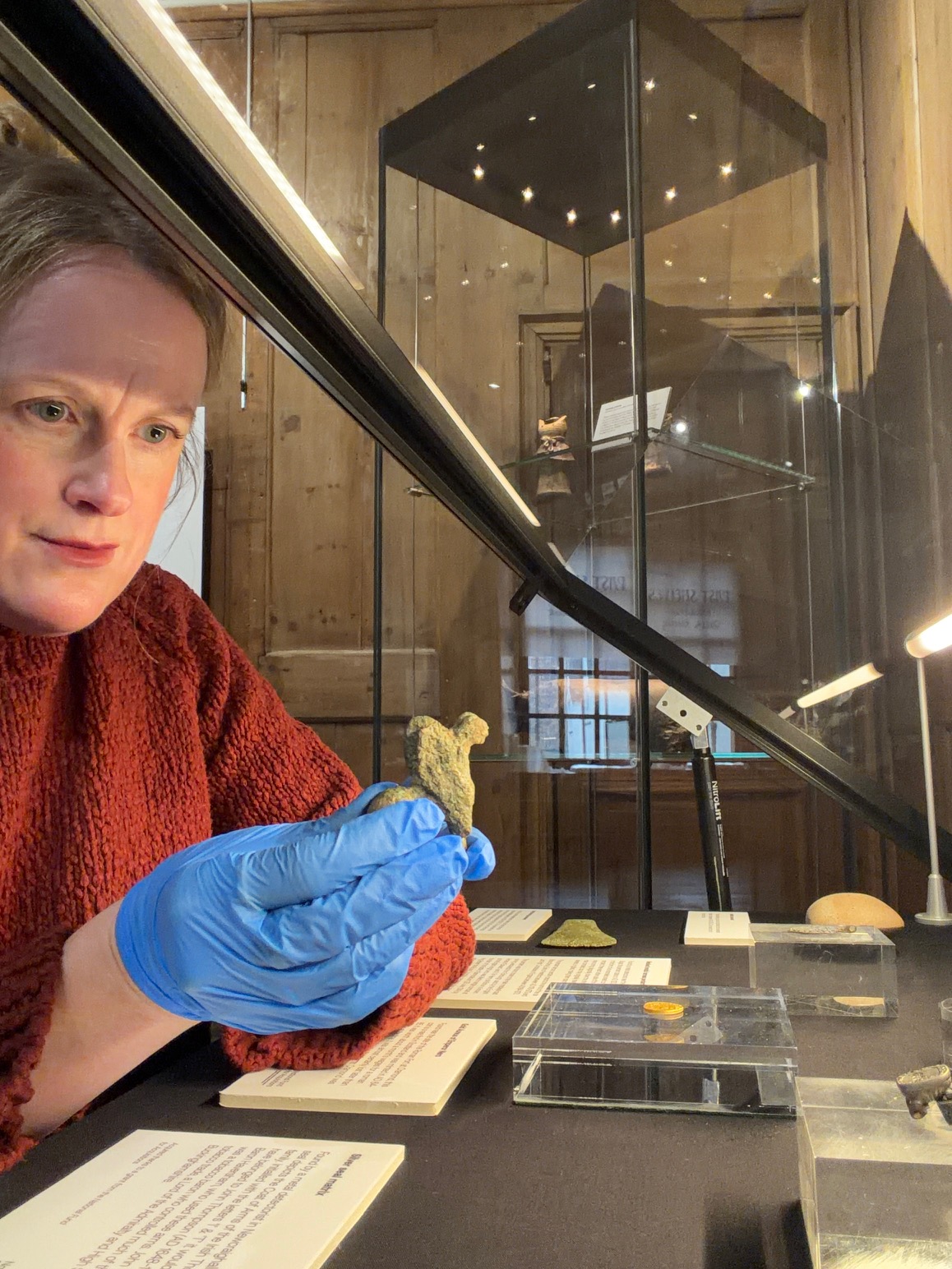 Photo of a woman in a red jumper wearing blue disposable gloves and holding a small carved stone object in a museum